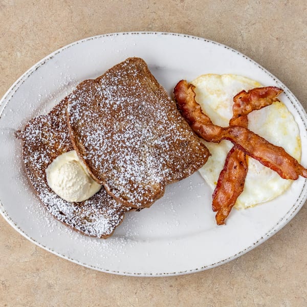 French toast with powdered sugar and whipped cream, served with bacon and fried eggs.