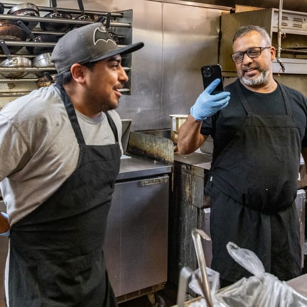 Two Chicken Run chefs in aprons, one taking a selfie with the other in a busy kitchen.
