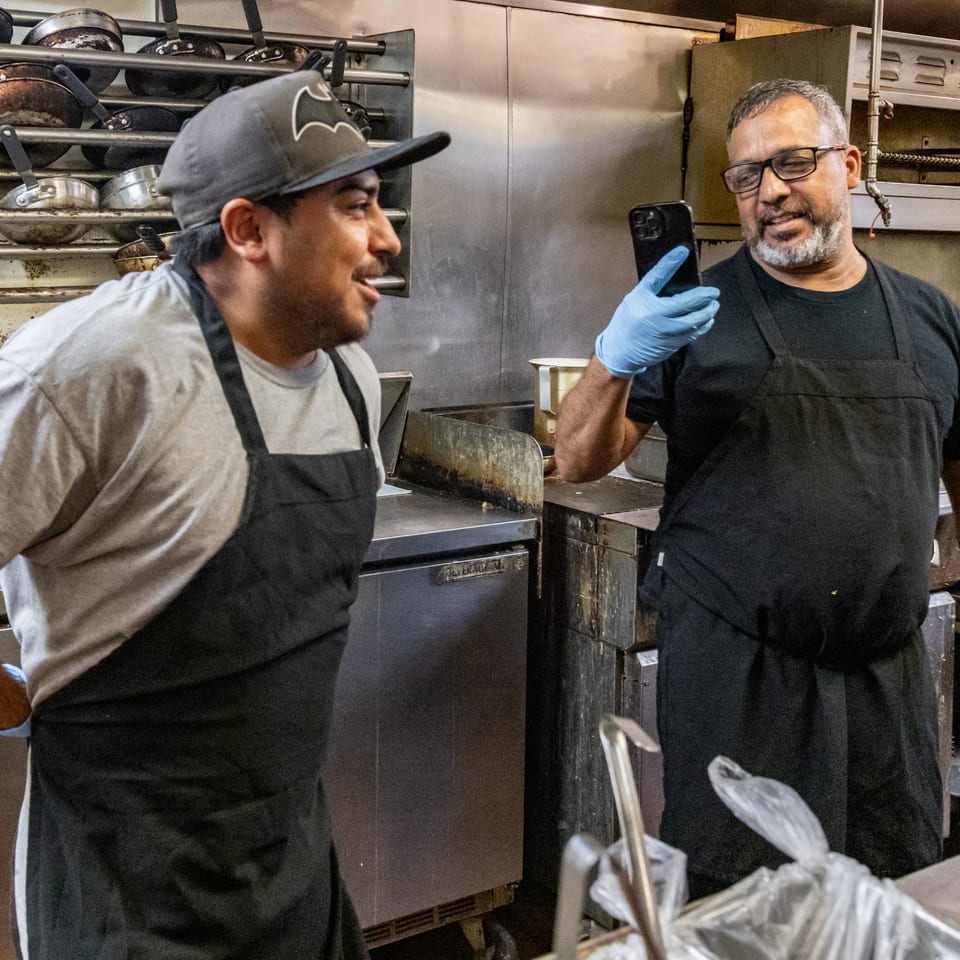 Two Chicken Run chefs in aprons, one taking a selfie with the other in a busy kitchen.