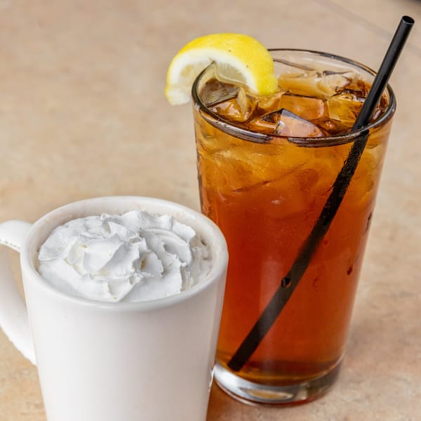 A white mug of hot chocolate with whipped cream next to a glass of iced tea with a lemon slice.