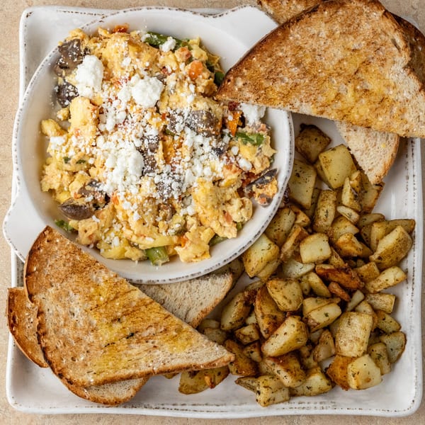 Scrambled eggs with feta, mushrooms, and vegetables, served with toast and seasoned potatoes.