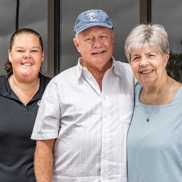 Three smiling people, two older and one younger, stand together in front of a building.