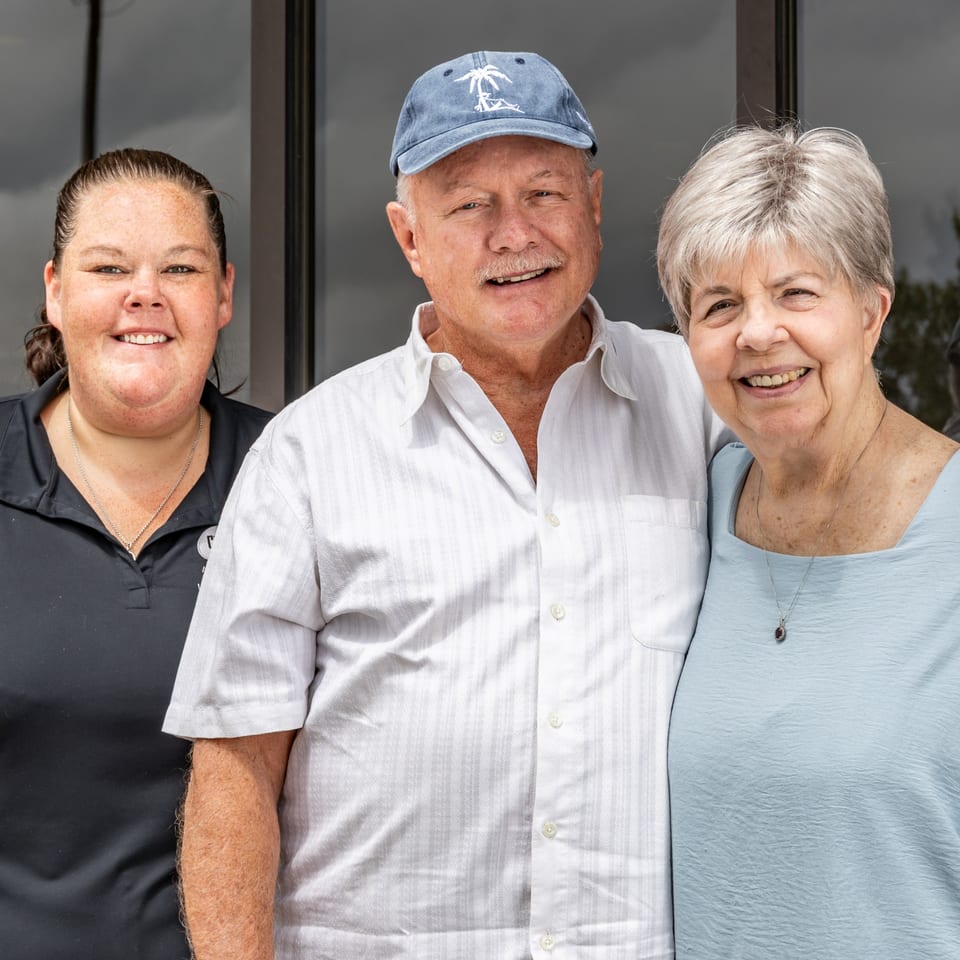 Three smiling people, two older and one younger, stand together in front of a building.