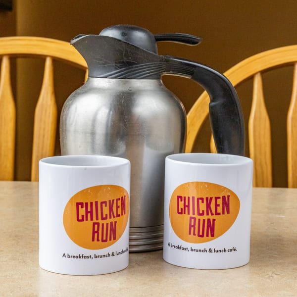 Two white Chicken Run mugs and a silver coffee carafe on a table with wooden chairs.