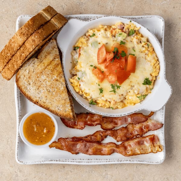 A breakfast skillet with melted cheese, tomatoes, and parsley, served with toast, bacon, and sauce.