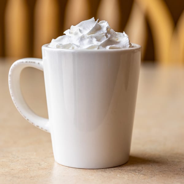 A white mug filled with whipped cream sits on a light brown table at Chicken Run.