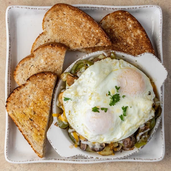 Breakfast skillet with fried eggs, potatoes, peppers, mushrooms, cheese, and a side of toast.